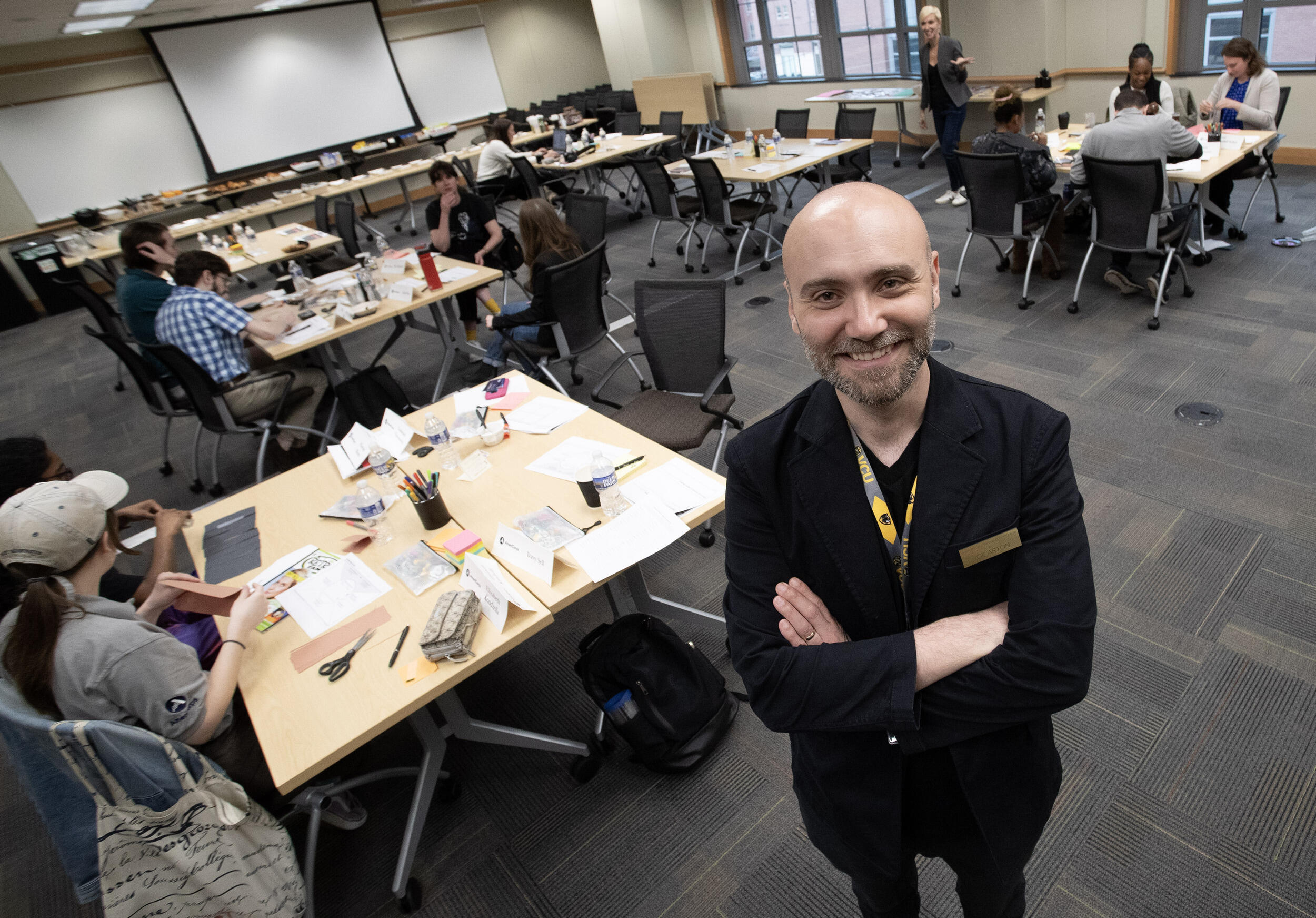 A photo of a man standing and smiling with his arms crossed against his chest. He is standing in a room full of tables with children sitting at them. 