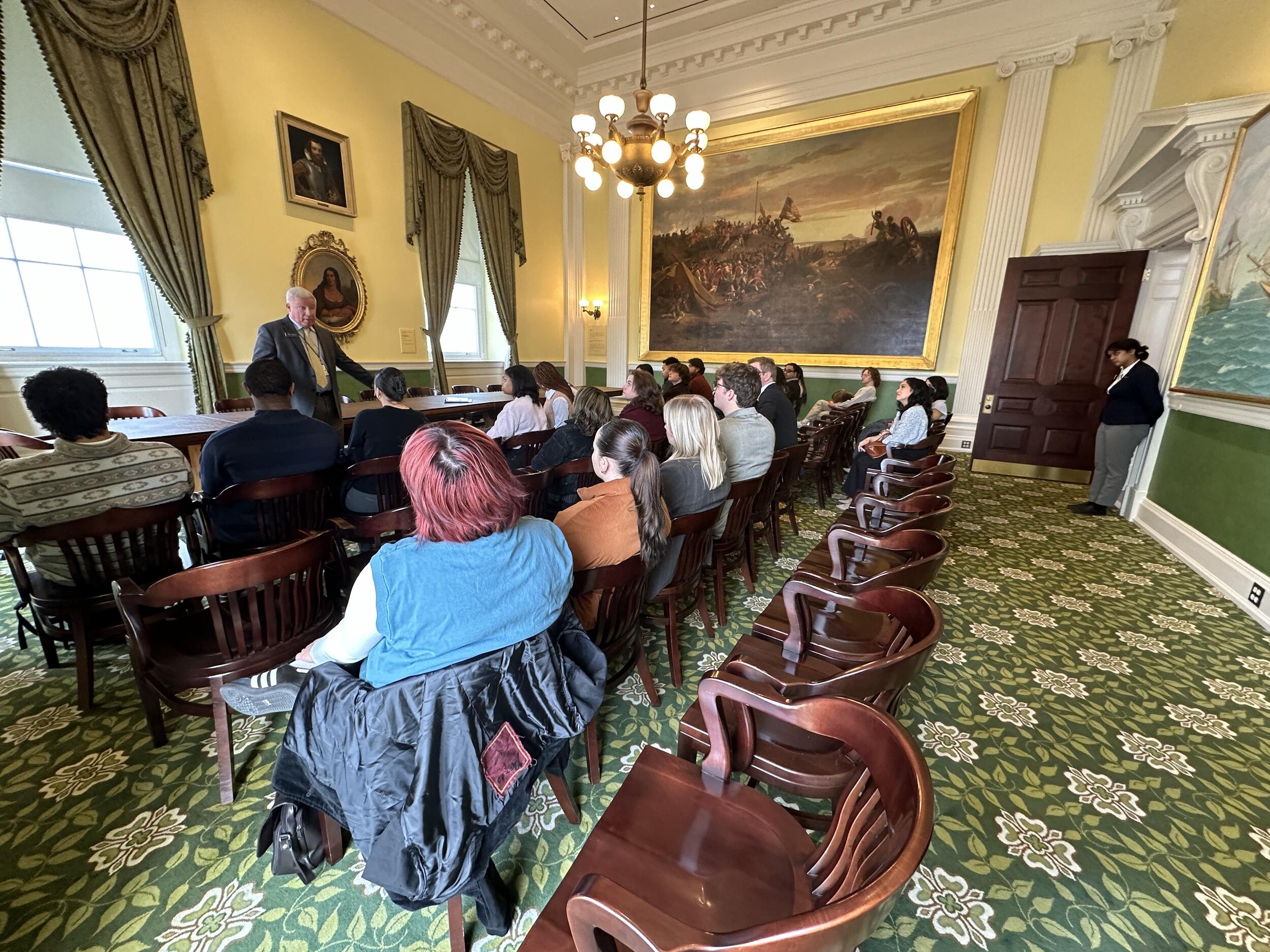 A photo of twenty two people sitting in chairs watching a man speak in the front of the room. At the back of the room a woman is standing against a wall. The walls of the room are yellow and the carpet is green. 