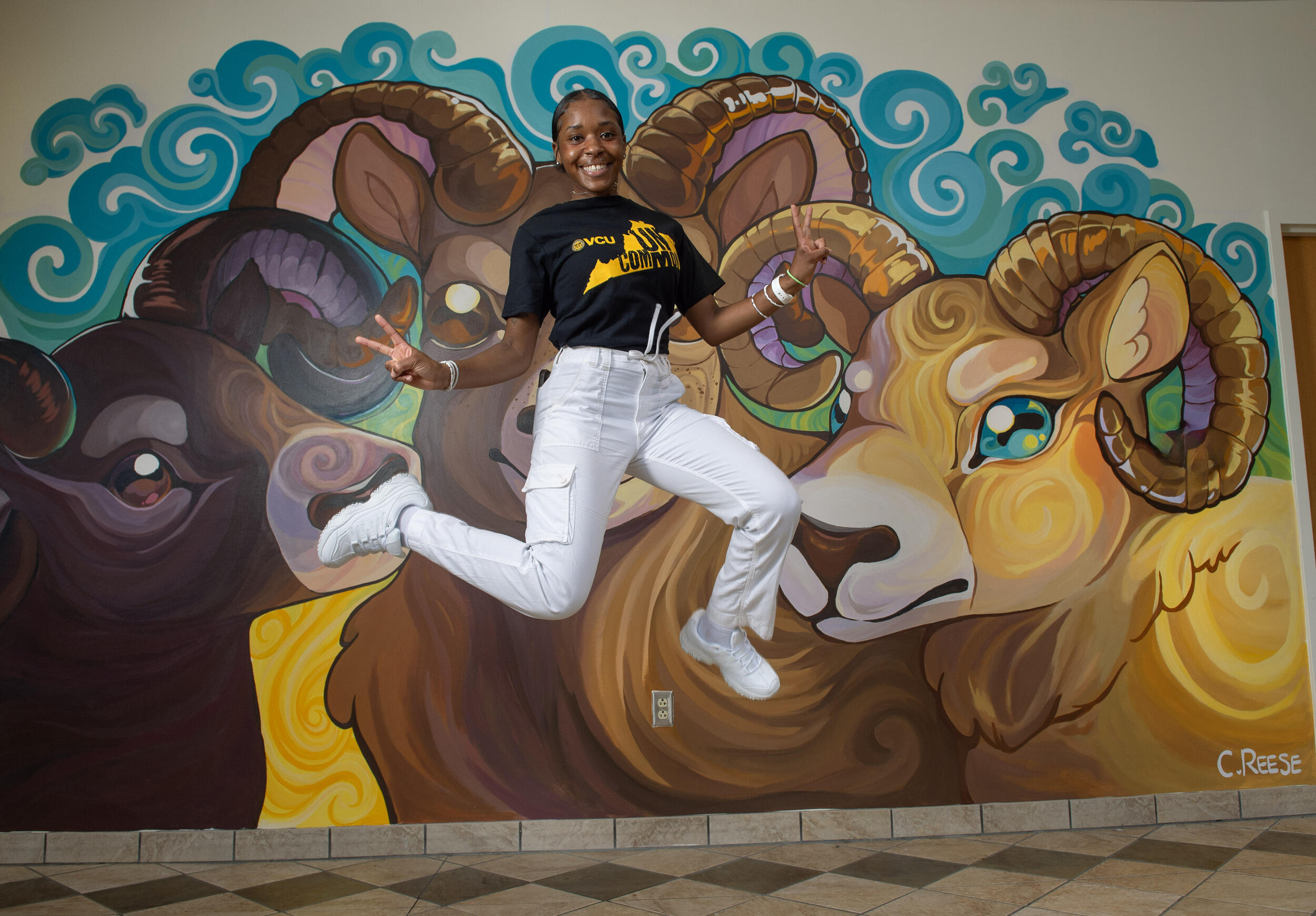 A photo of a woman wearing a black thisrit and white pants jumping. Both of her hands are making peace signs. Her t-shirt has the shape of Virginia printed on it. Inside the state's silhouette is black text that reads \"UNCOMMON.\" Behind the woman is a mural of three rams. 