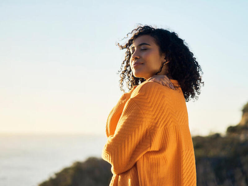 A woman wearing a yellow sweater standing on a cliff side next to the water. She is looking to the left. 