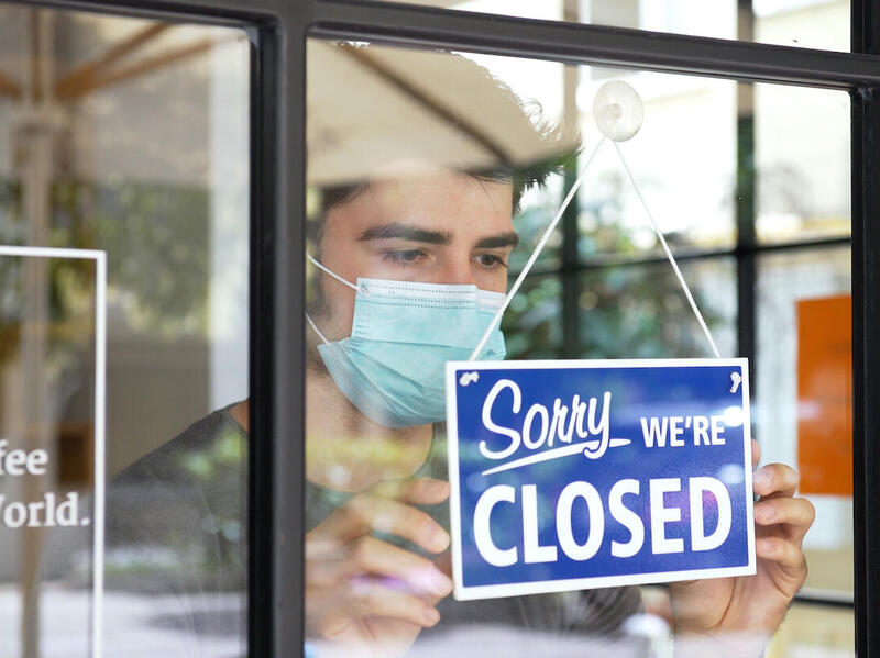 A person wearing a mask holds a \"Sorry we're closed\" sign attached on a window.