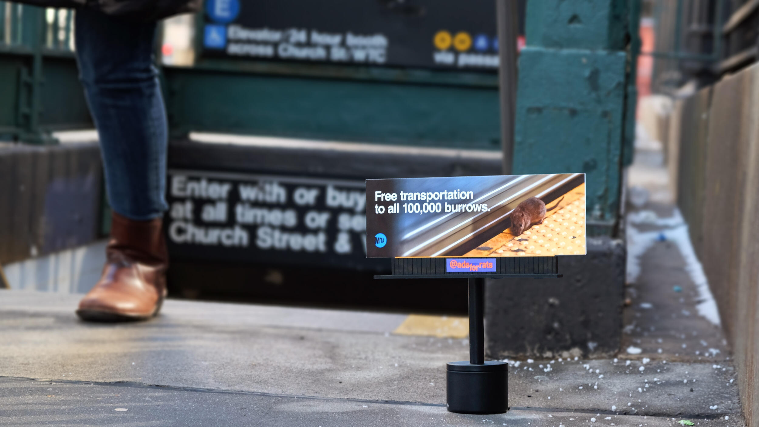 A photo of a mini billboard with the image of a rat on a train platform. White text reads \"Free transportation to all 100,000 burrows.\" \"MTA\"