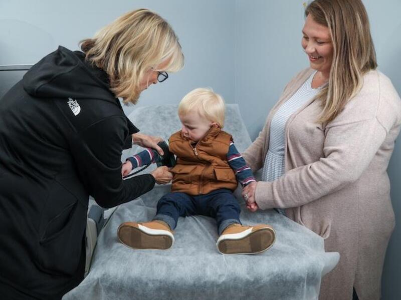 A photo of a todler sitting on a medical examination table. On the left and right of the table are two women looking at the baby. 