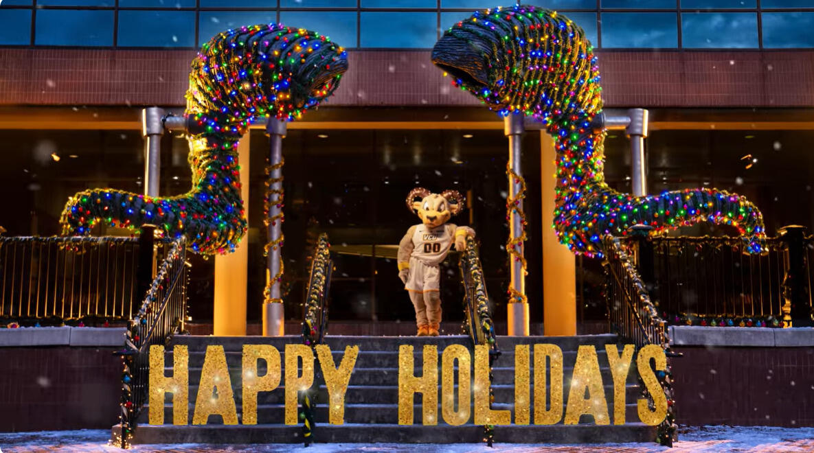 A photo of Rodney the Ram standing under a statue of ram horns that is covered in tinsel and Christmas lights. There is gold text under him that read \"HAPPY HOLIDAYS.\"