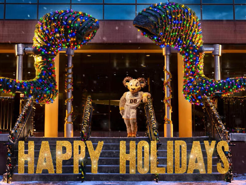 A photo of Rodney the Ram standing under a statue of ram horns that is covered in tinsel and Christmas lights. There is gold text under him that read \"HAPPY HOLIDAYS.\"