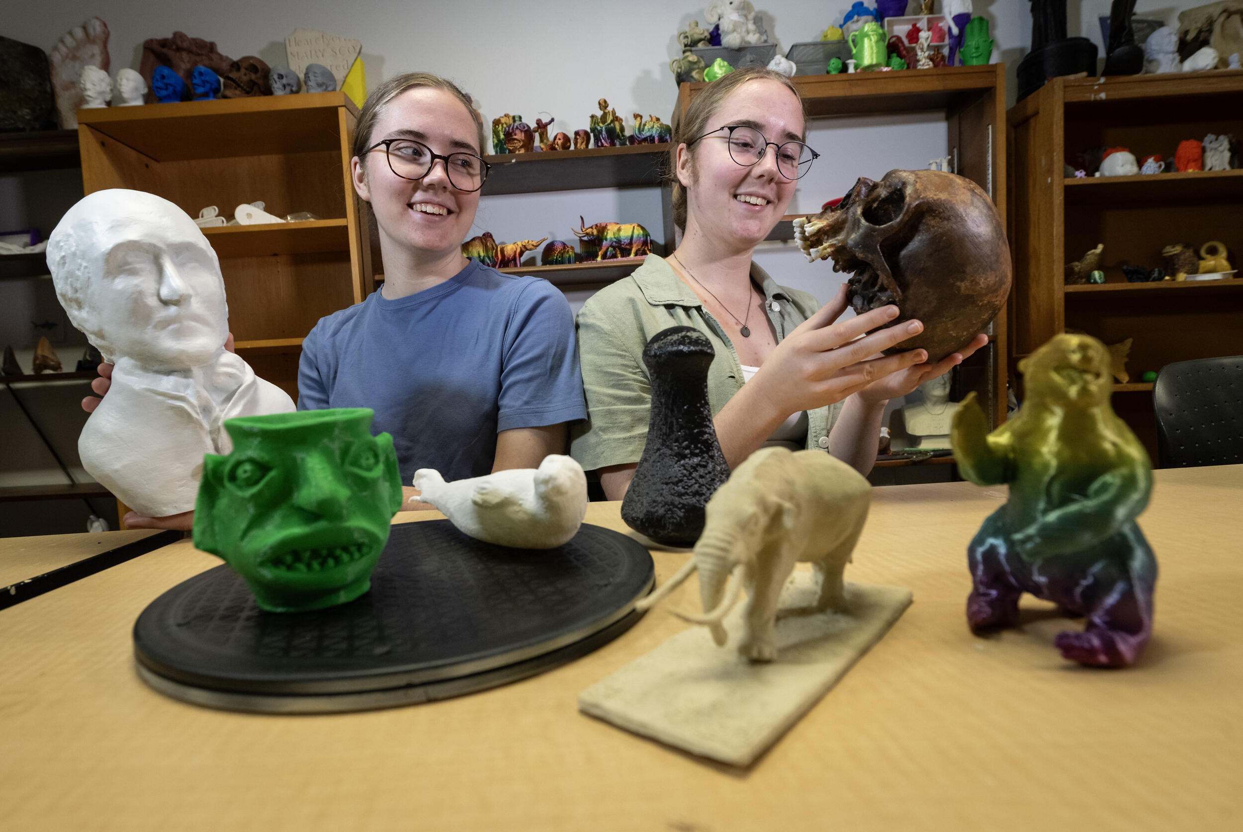 A photo of two women sitting at a table covered in objects. 