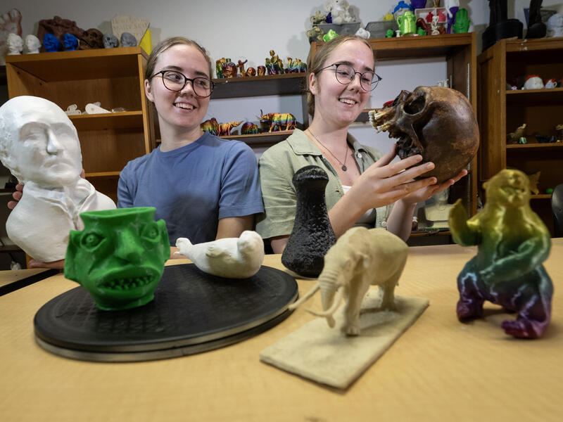 A photo of two women sitting at a table covered in objects. 