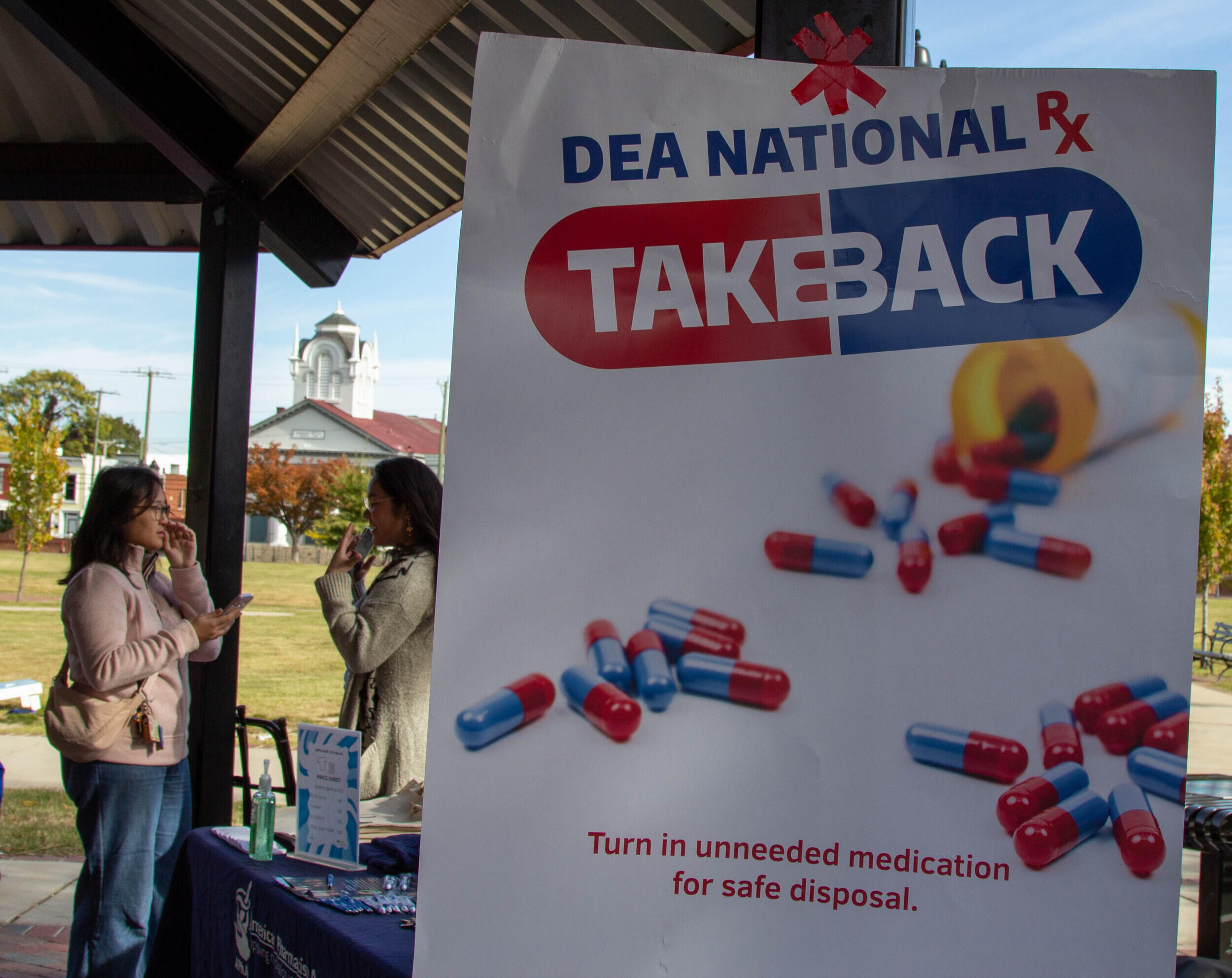 A photo of two women talking to each other next to a table. In front of them is a poster that says \"DEA NATIONAL TAKEBACK\" \"Turn in unneeded medication for safe disposal.\" with a photo of an open pill bottle on it. 