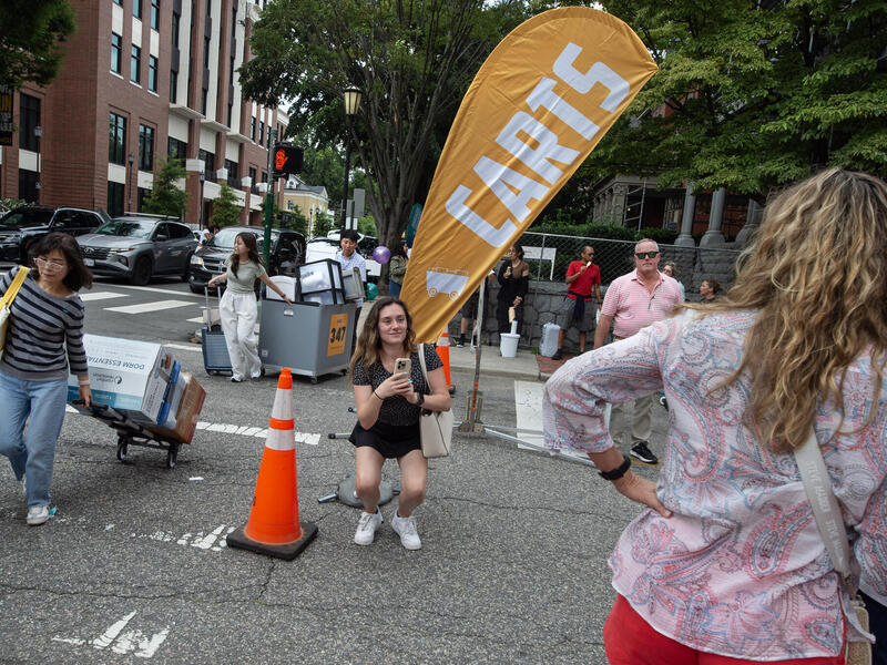 A photo of a street with people walking across it carrying luggage and bins filled with boxes. There is a person taking a photo of two other people. 