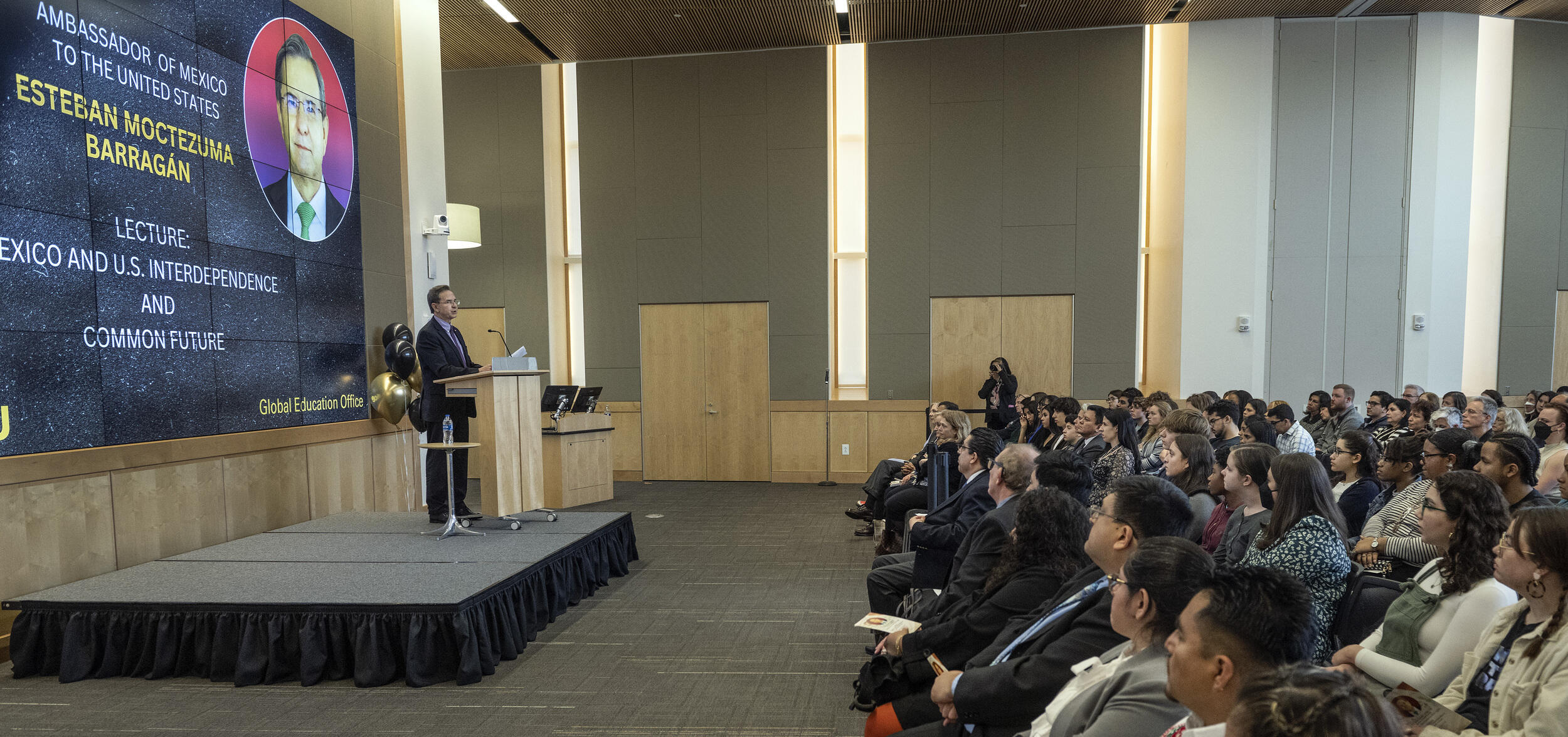 A photo of a man standing at a podium in front of a room filled with people sitting in chairs. 