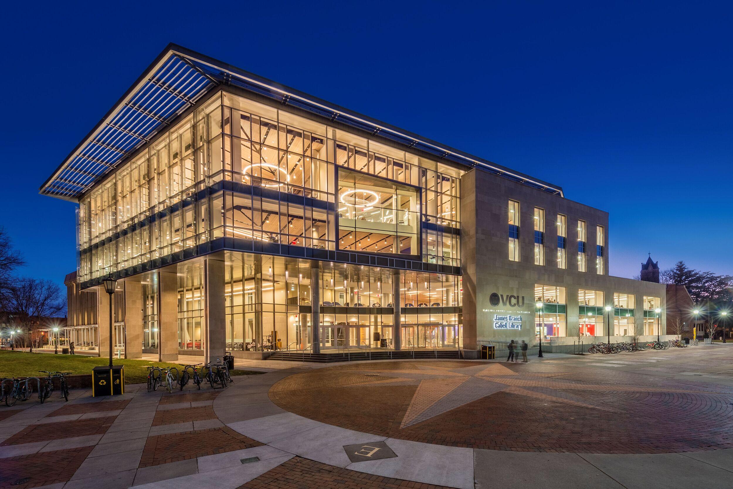 A photo of James Branch Cabell Library at dawn. The sky is a dark blue and the entrance to the library is lit up. 