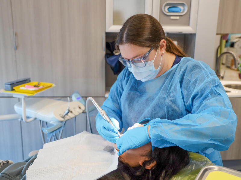 A photo of a woman wearing blue scrubs, gloves, glasses and a face mask cleaning the teeth of a person lying down. The person lying down is wearing a white disposable bib. 