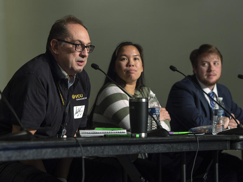 A man, woman, and another man sit at a table with microphones in front of them. THe man on the far left is speaking and the other two people at the table are looking at him. 