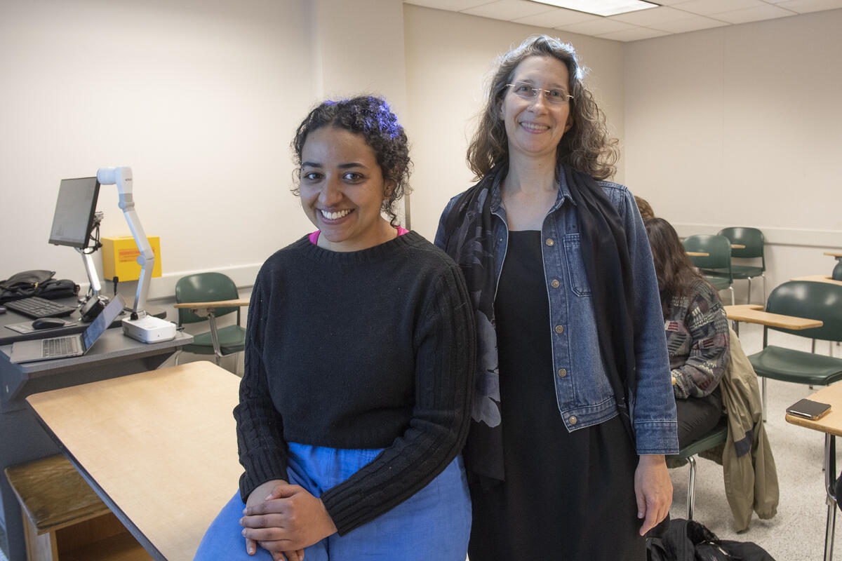 Two women in a classroom. The woman on the left is sitting on a table and the woman on the right is standing next to her. There are desks behind them. 