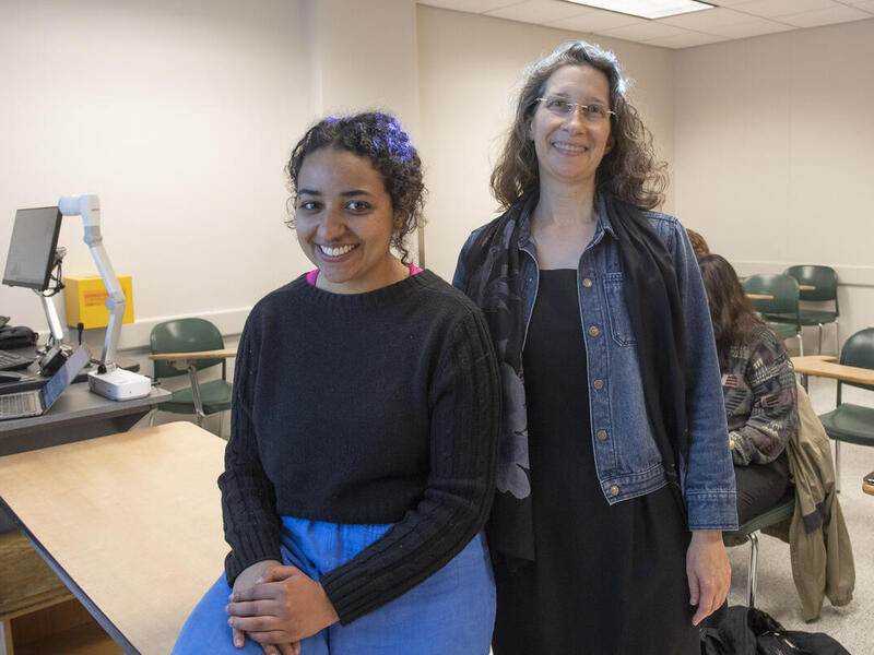 Two women in a classroom. The woman on the left is sitting on a table and the woman on the right is standing next to her. There are desks behind them. 