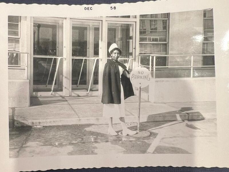 A black and white photo of a woman wearing a nurse uniform in front of a building 