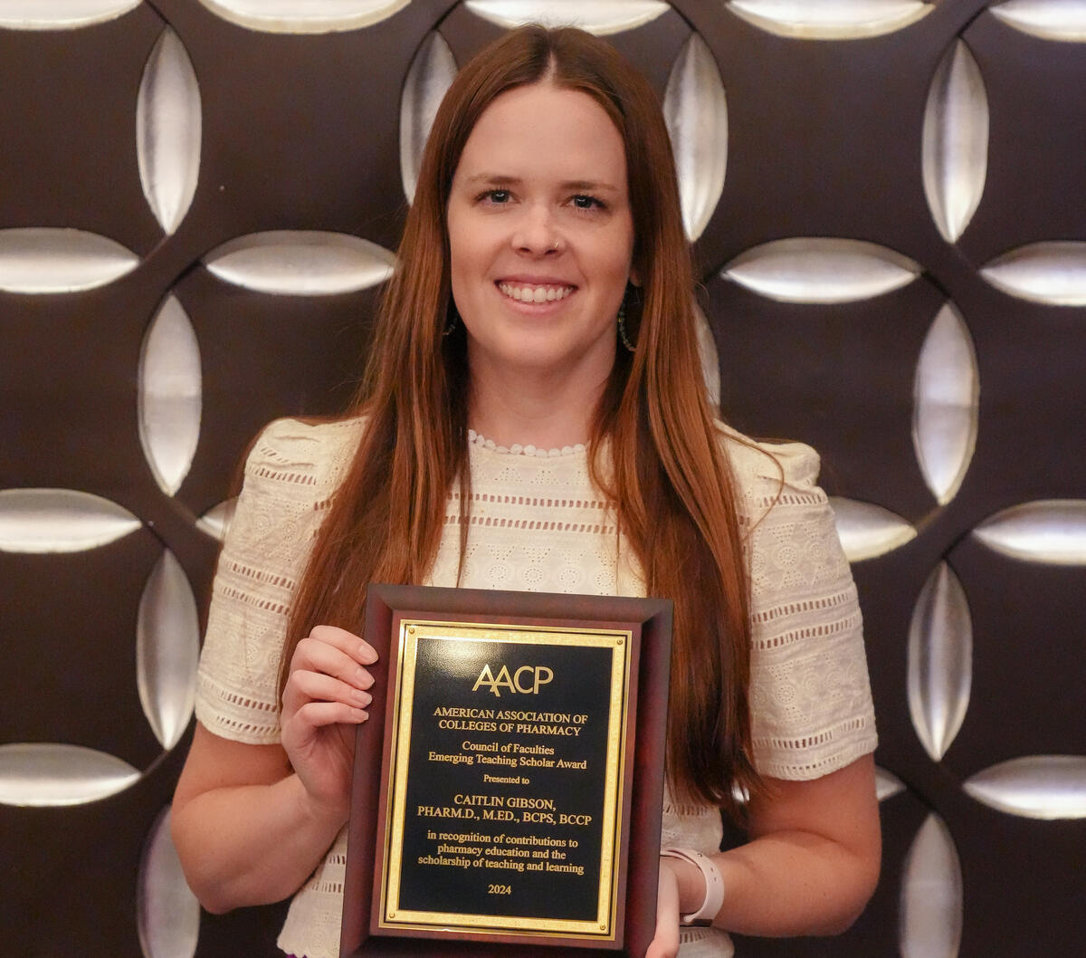 A photo of a woman holding a plaque with gold letters. The plaque reads \"AACP AMERICAN ASSOCIATION OF COLLEGES OF PHARMACY\" \"Council of Faculties Emerging Teacher Scholor Award\" \"Presented to CAIT GIBSON, PHARM.M.D., BCPS, BCCP\" \"in recognition of contributions to pharmacy education and the scholarship of teaching and learning\" \"2024\"