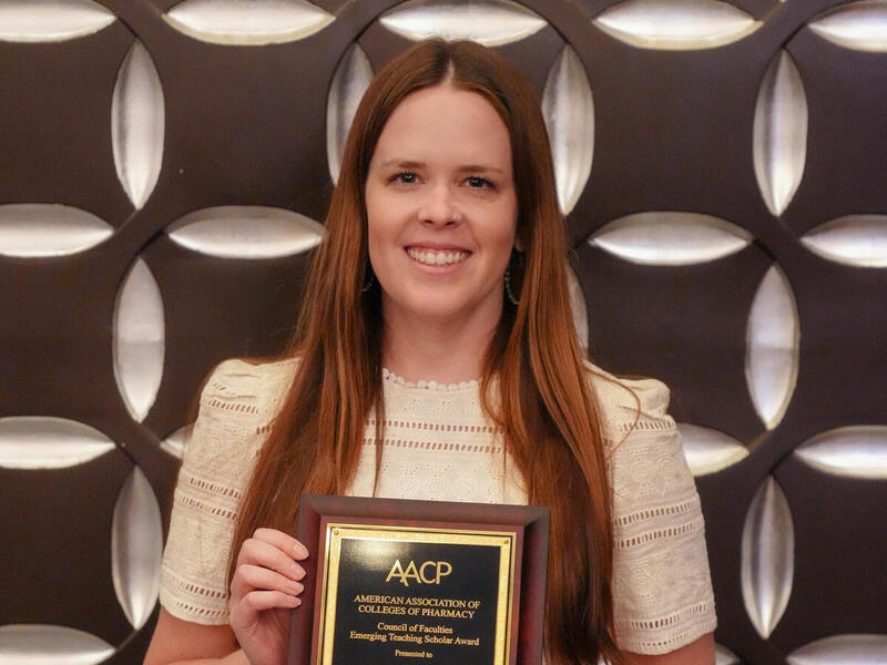 A photo of a woman holding a plaque with gold letters. The plaque reads \"AACP AMERICAN ASSOCIATION OF COLLEGES OF PHARMACY\" \"Council of Faculties Emerging Teacher Scholor Award\" \"Presented to CAIT GIBSON, PHARM.M.D., BCPS, BCCP\" \"in recognition of contributions to pharmacy education and the scholarship of teaching and learning\" \"2024\"