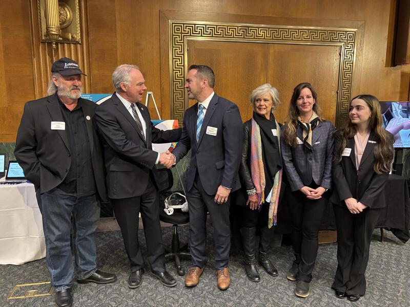 A group photo of six people standing. The second and third man on the left are facing each other and shaking hands. 