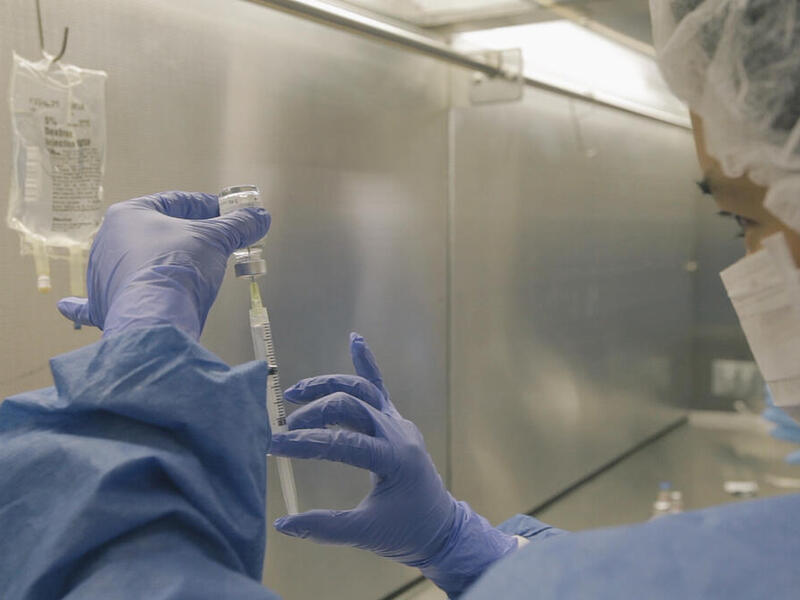 A photo of a person wearing blue gloves, a white face mask, a white hair net, and blue disposable medical scrubs filling a syringe. 