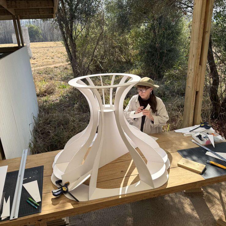 A woman works at a work table outdoors on a large piece of a sculpture.