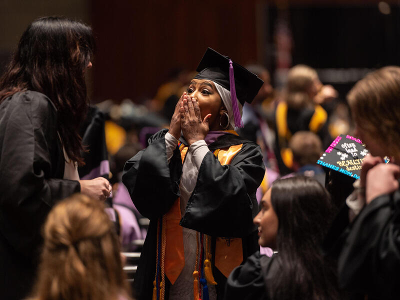 A student in cap and gown holds her hands to her face in excitement. 