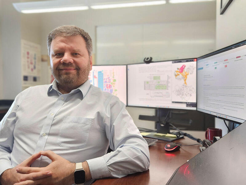 A photo of a man from the waist up. He is sitting at a desk that has three computer monitors on it. 