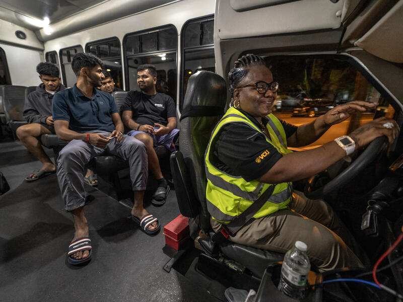 A photo of a woman driving a vehicle. Behind her are four men sitting in the rear passenger seats. 