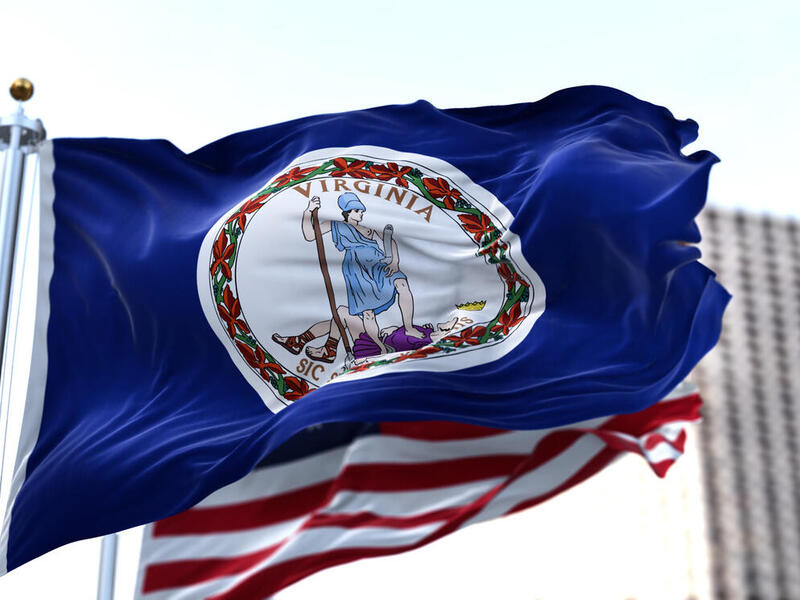 A photo of the Virginia State flag flying in front of the American flag. Behind the two flags is a high-rise building. 