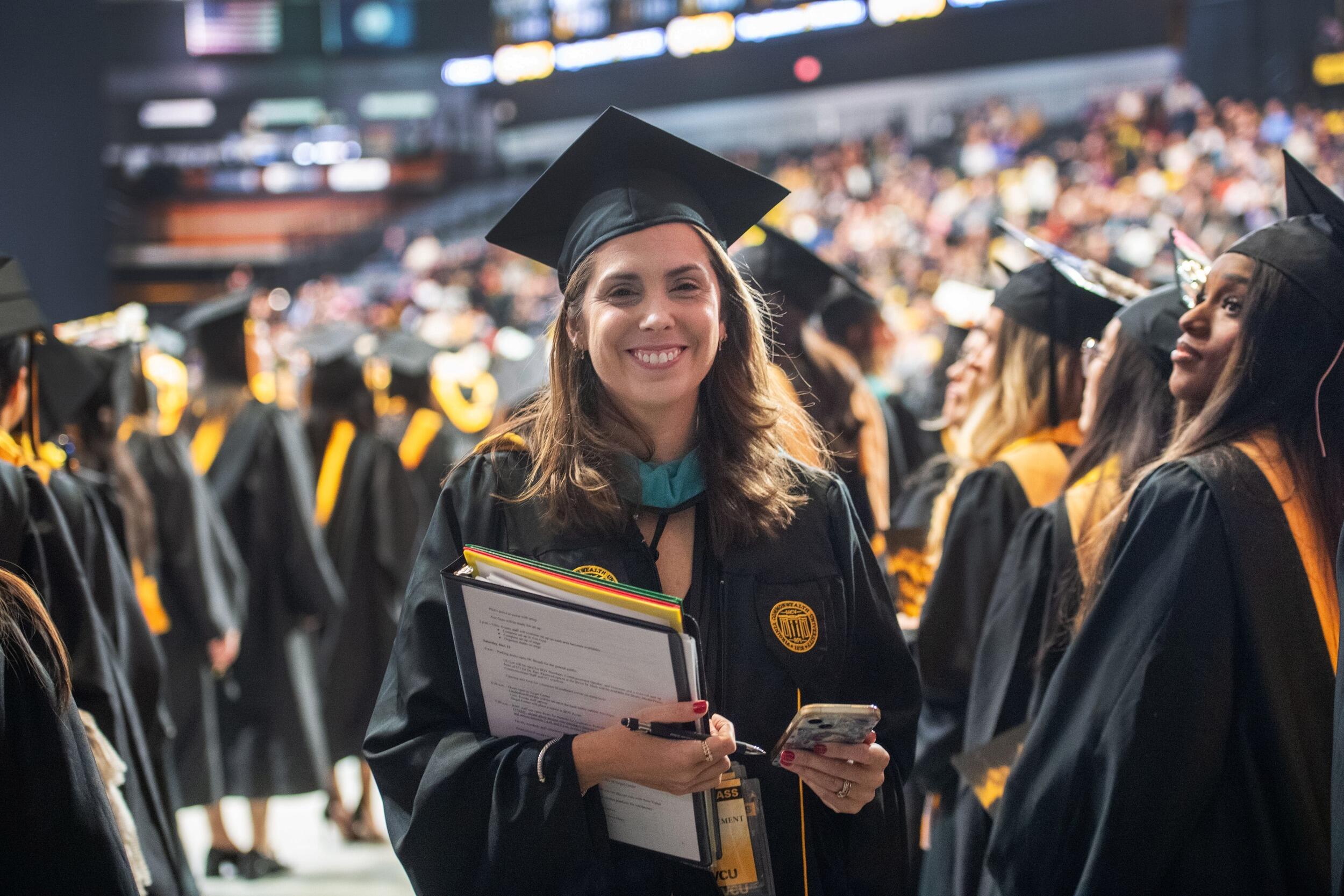 A photo of a woman wearing a cap and gown smiling and standing. In her left hand she has a folder and a binder. In her right hand she is holding a cell phone. All around her is a crowd other people in graduation caps and gowns. 