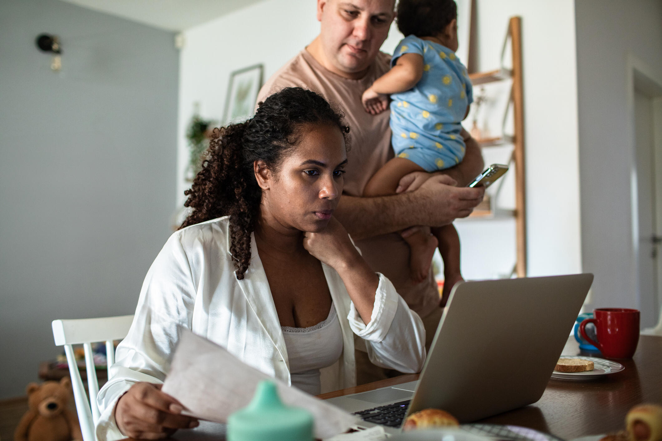 A photo of a woman sitting at a table looking at a laptop and holding a piece of paper. Next to her is a man holding a baby in his arms and also looking at the laptop.