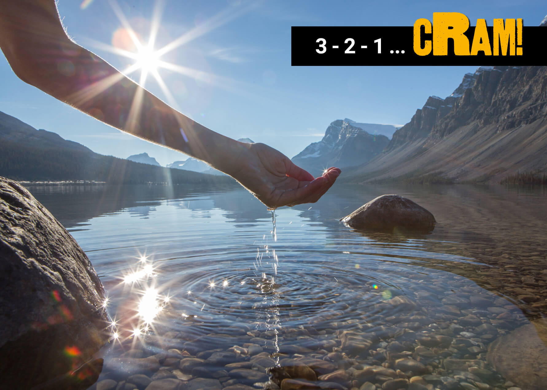 A photo of a hand scooping up water from a clear lake. The botoom of the lake is covered in small rocks, and two big rocks are sticking out of the water. Behind the lake are mountains. 
