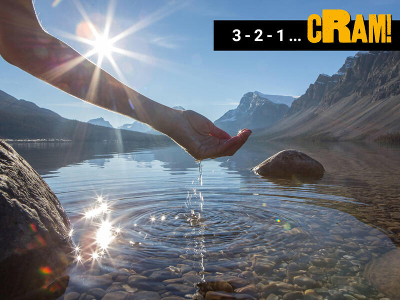 A photo of a hand scooping up water from a clear lake. The botoom of the lake is covered in small rocks, and two big rocks are sticking out of the water. Behind the lake are mountains. 