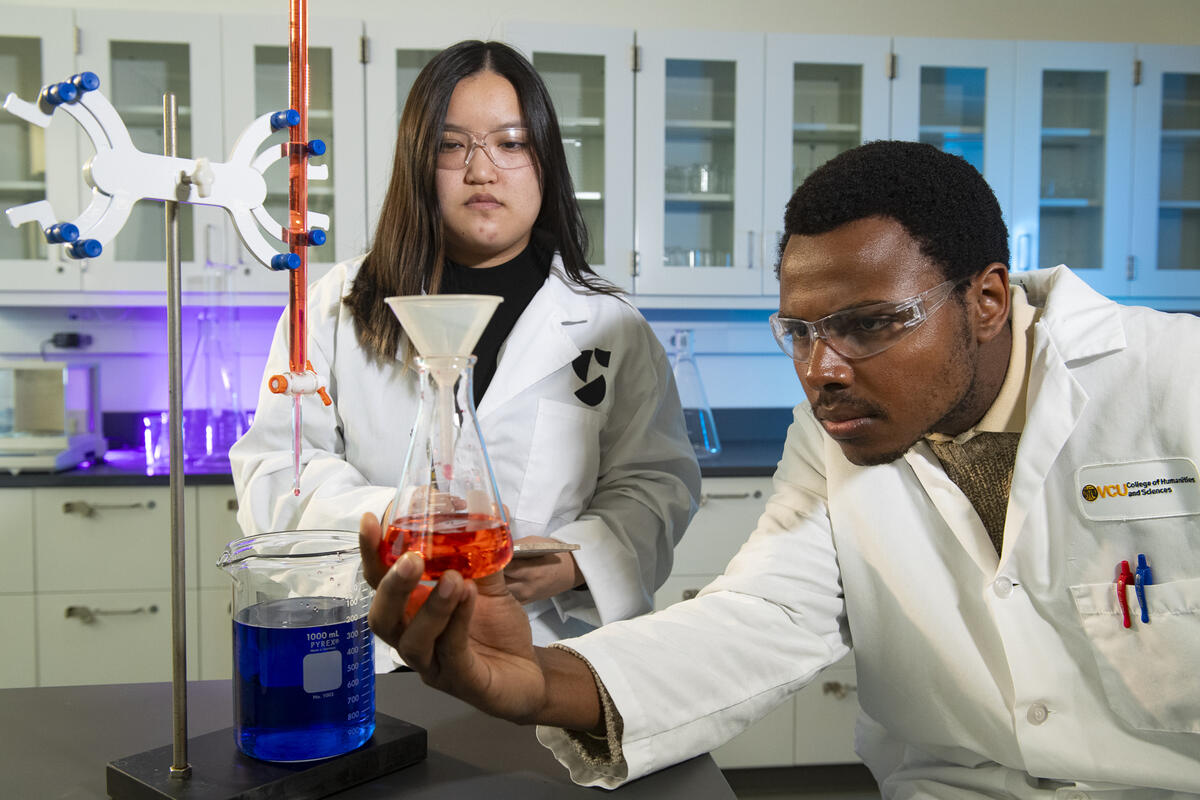 A photo of a man in a white libe coat holding a glass beaker with a red liquid in it. Next to him, a woman in a white labcoat is standing and looking at the bottle. 