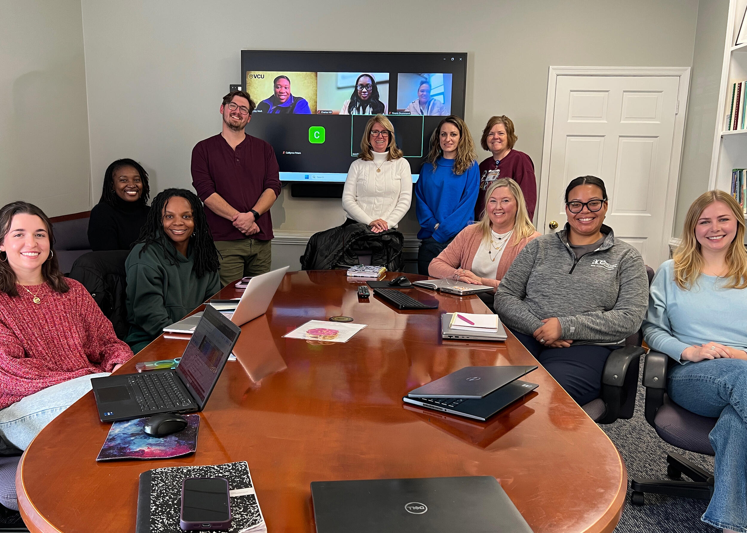 A photo of 10 people in a room around a large table and three people being shown on a screen behind them. 