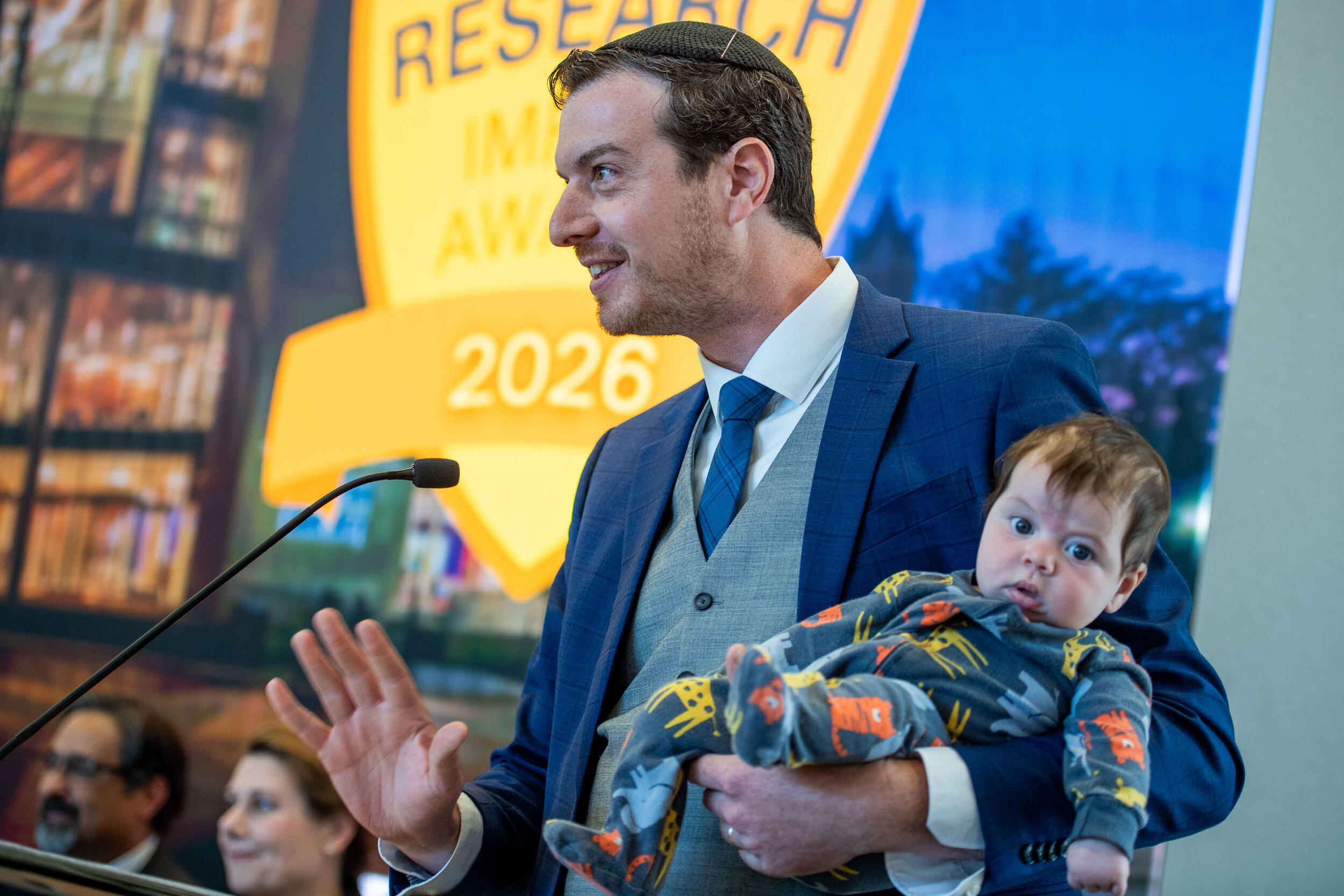 A photo of a man standing behind a podium and speaking while holding a baby. 