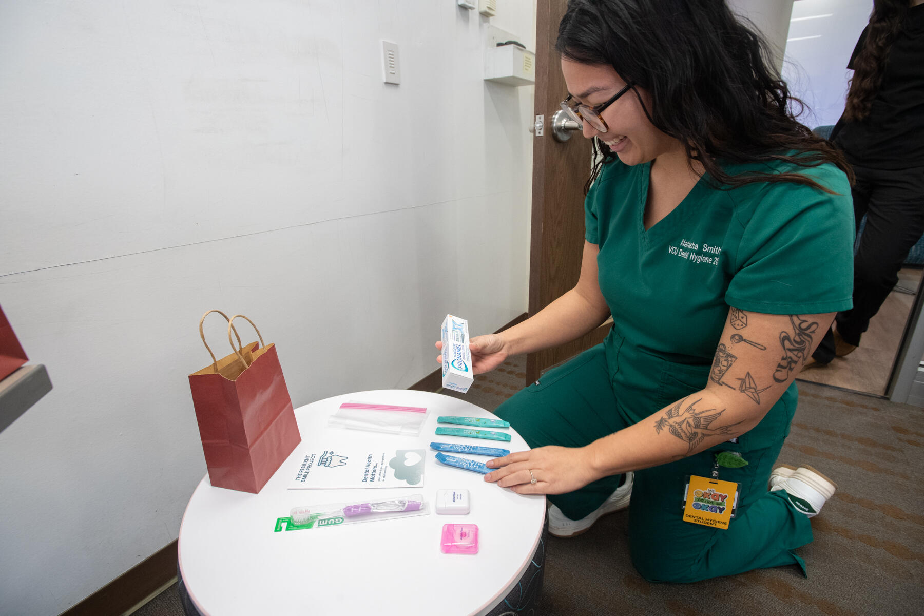A photo of a woman in scrubs kneeling next to a white circular table and placing dental hygiene products on it. 