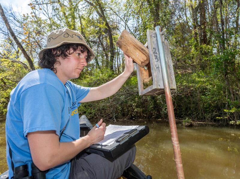 A photo of a woman writing on a clip cloard and looking in a wooden bird house 