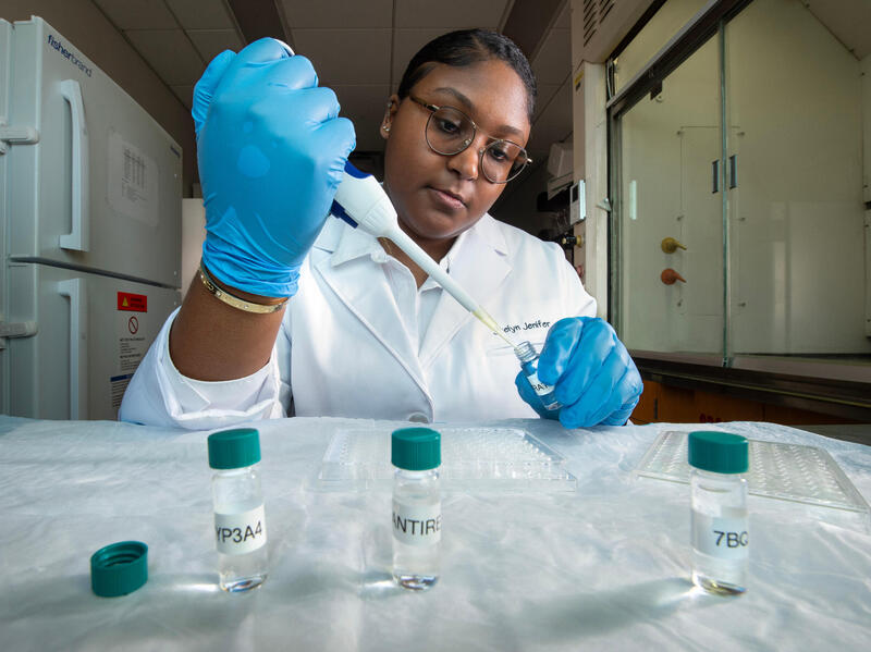 Student in lab coat and gloves uses a pipette.