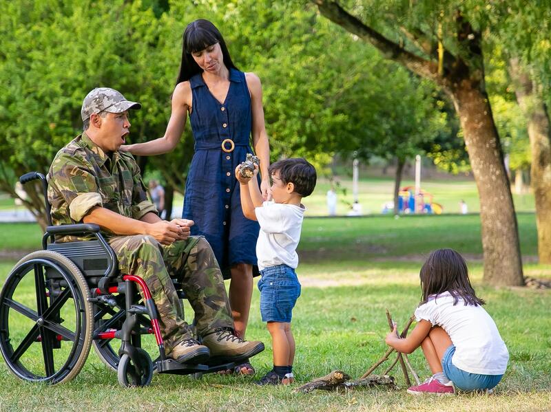 A photo of a man sitting in a wheel chair with a woman standing to his right with her hand on his shoulder. He is looking at a small boy in front of him who is holding up a large stick. Behind the boy is a small girl sitting on the ground playing with other sticks. 