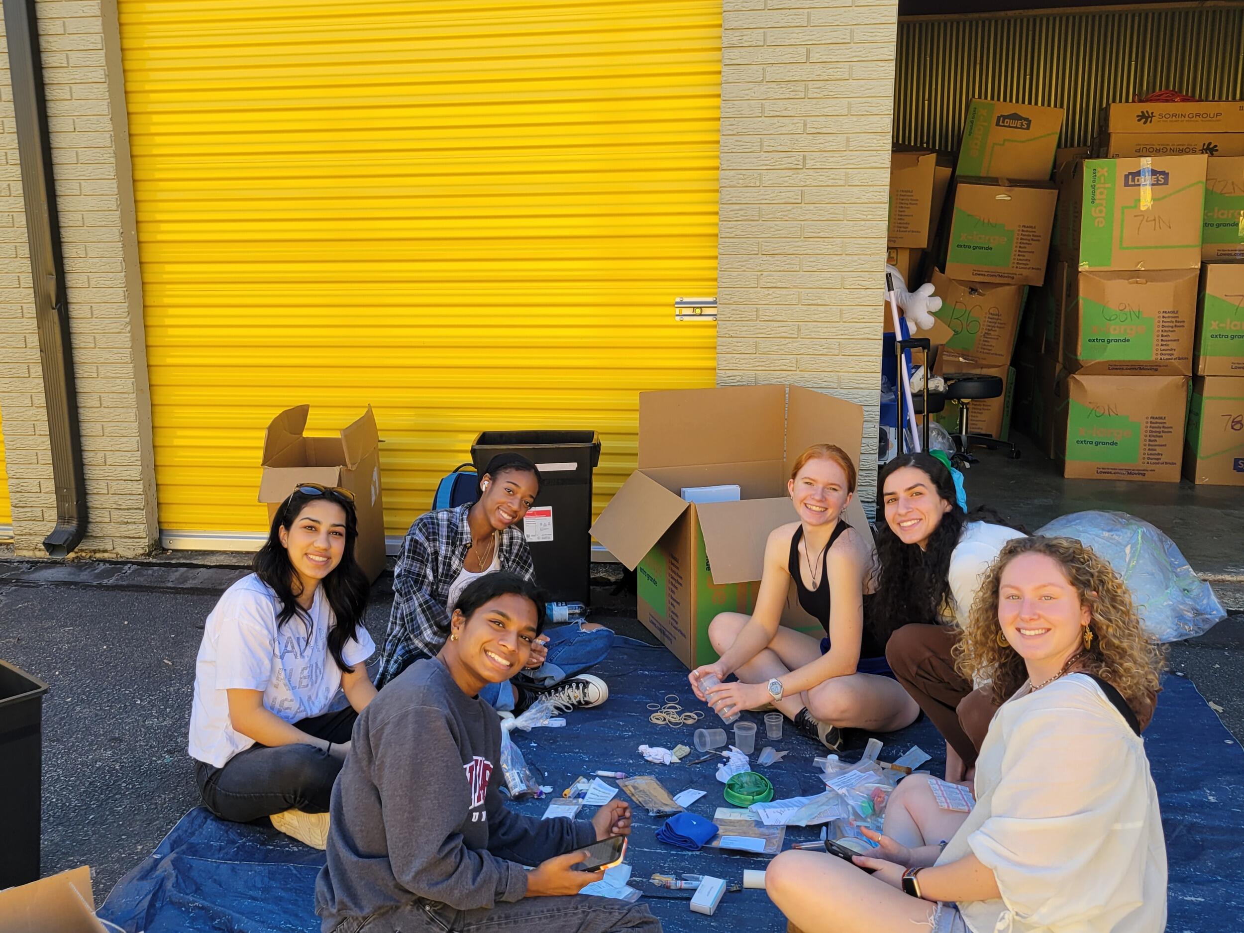 A group of students sitting on a blue tarp in a circle. They are sitting in front of storage units, one with its door closed and the other open and full of boxes. 