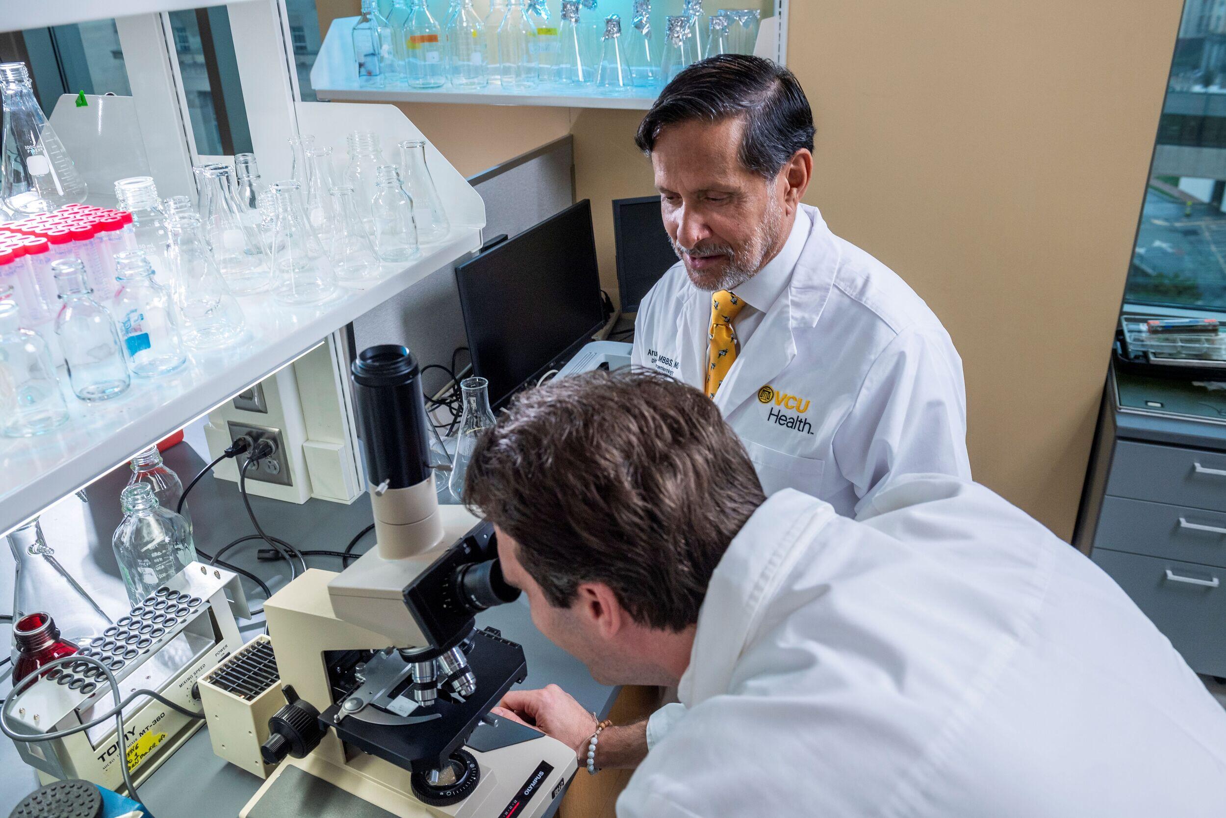 A man looks through a microscope while a man nearby in a lab coat watches him.