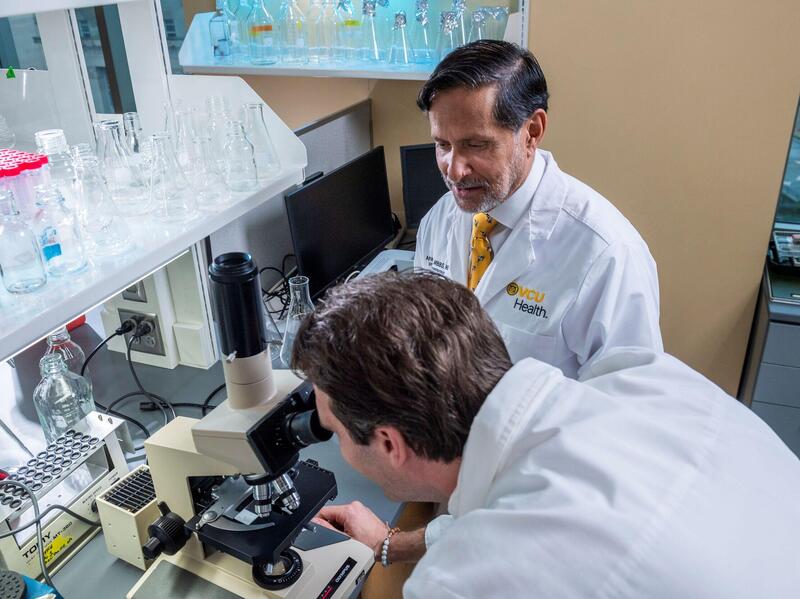 A man looks through a microscope while a man nearby in a lab coat watches him.