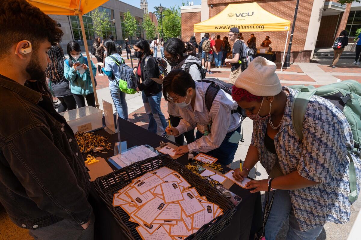 People standing under a tent around a table. Two people are writing on pieces of paper and a person on the other side of the table is watching them. 