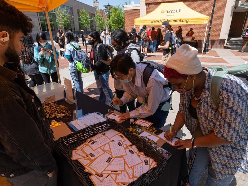 People standing under a tent around a table. Two people are writing on pieces of paper and a person on the other side of the table is watching them. 