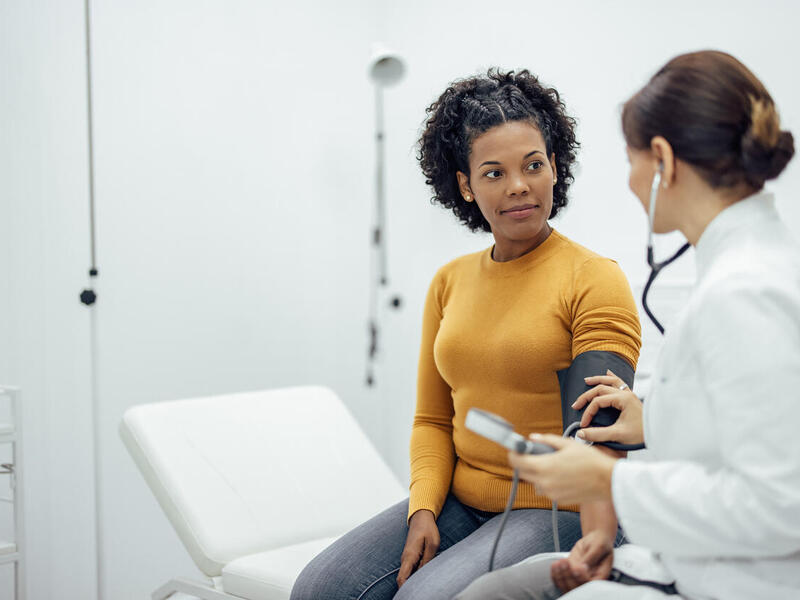A photo of a woman sitting on an examination table at a doctor's office with a blood pressure band around her arm. Next to her is a doctor who had a stethoscope on who is listening to the women's hearbeat through the band. 