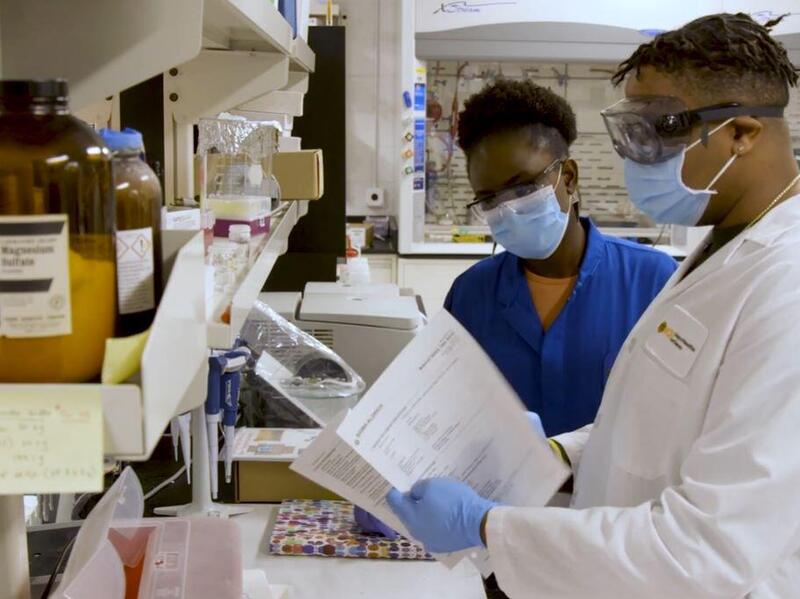 Two people wearing lab coats, safety glasses, and face masks standing in a laboratory 