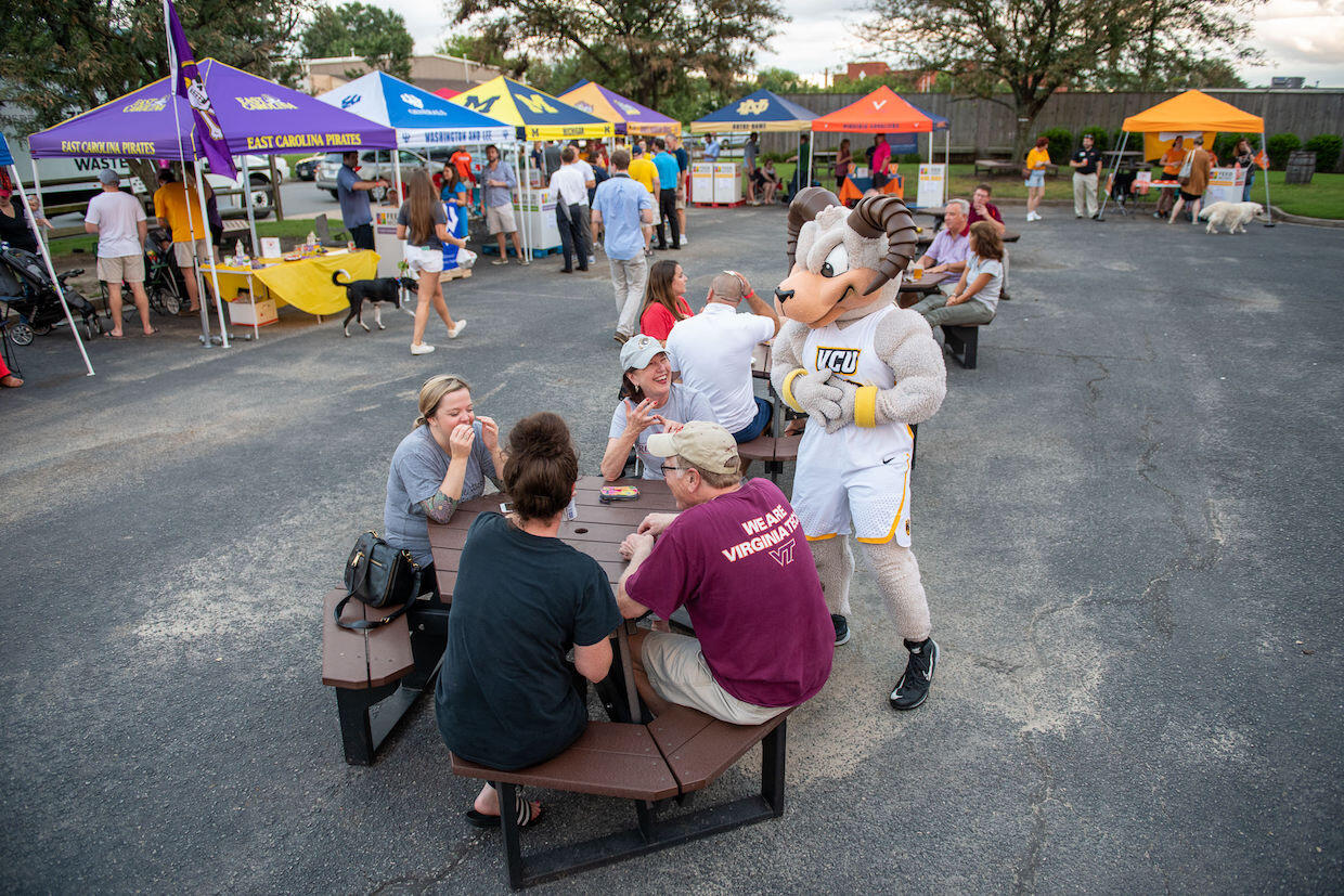 Rodney the Ram interacts with a group of people sitting at a picnic table. Tents representing various universities - East Carolina University, Washington and Lee University, University of Michigan, James Madison University, University of Notre Dame, University of Virginia, and the University of Tennessee - are set up in the background.
