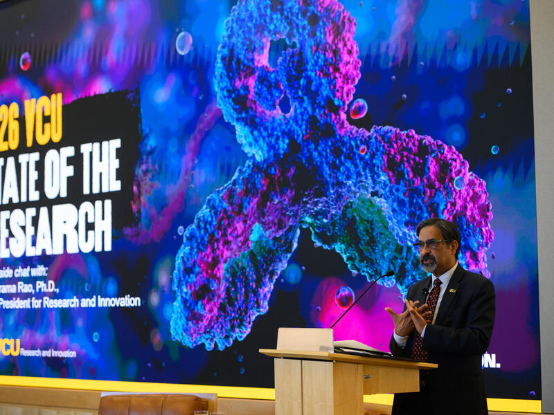 A photo of a man standing behind a podium and speaking. Behind the man is a screen that says \"2026 VCU STATE OF THE RESEARCH.\" 