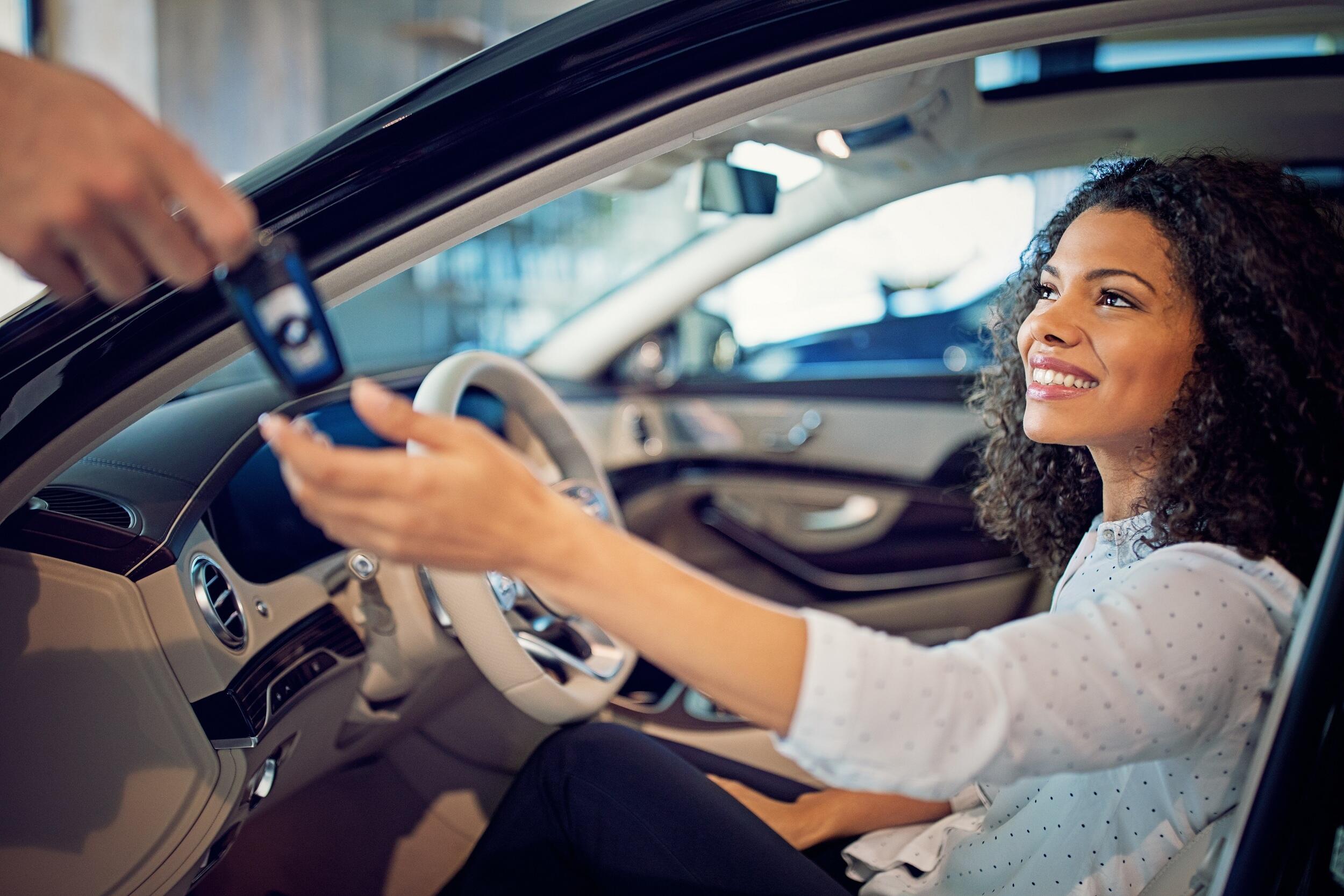 A woman sitting in a car with an open door smiling and reaching out to a hand giving her a car key